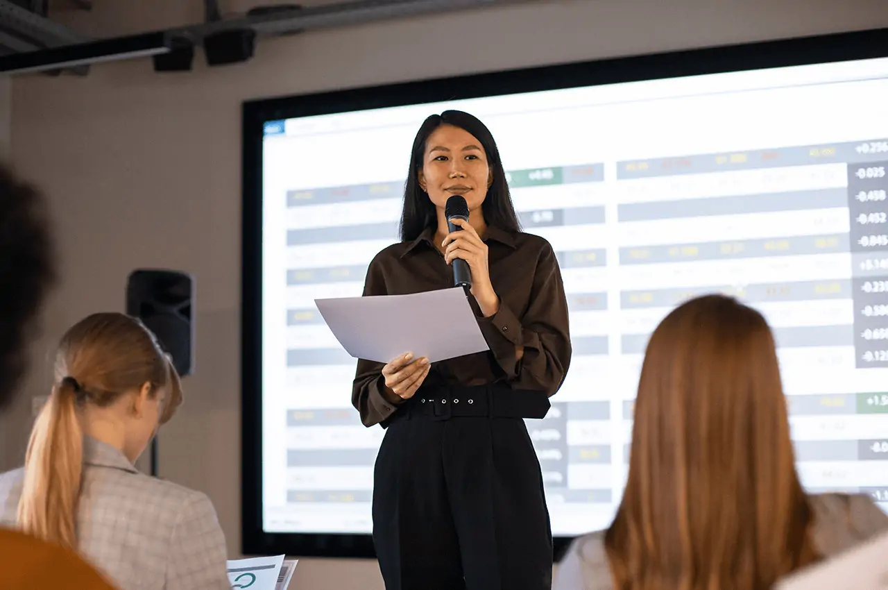 mulher de pé com microfone na mão e pessoas sentadas assistindo palestra em evento corporativo
