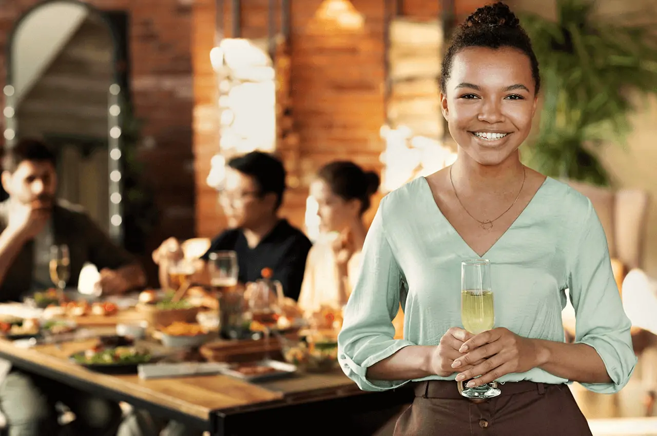 mulher de pé segurando uma taça de champanhe e ao fundo pessoas sentadas na mesa